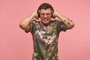 joven tatuado chico en de flores camiseta escuchando música también alto, cubierta orejas, bizco y mordiendo el bala, posando terminado rosado antecedentes foto