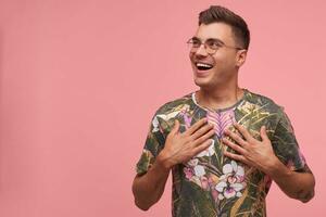 Portrait of cute young guy in flowered t-shirt, keeping palms on his chest, posing over pink background, looking away with wide smile photo