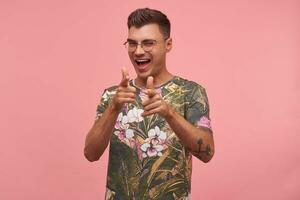 retrato de un alegre joven hombre, señalando a cámara, dando guiño y sonriente en general, aislado en rosado fondo, vistiendo de flores camiseta y lentes foto