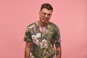 Shot of sad crying young man in glasses, wearing flowered t-shirt, looking unhappy and upset, standing over pink background, negative emotions concept photo