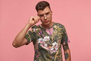 estudio Disparo de joven bonito chico con lánguido mirar, vistiendo camiseta con floral imprimir, conmovedor su anteojos, en pie terminado rosado antecedentes foto