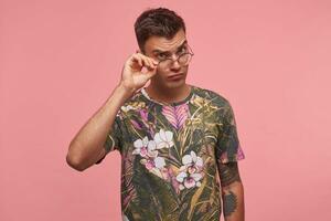 Attractive young guy taking off eyewear and looking from under forehead at camera with serious expression, posing over pink background photo