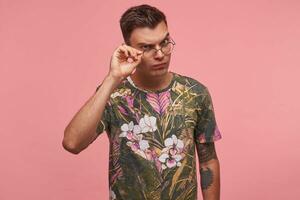 retrato de joven hombre en de flores camiseta con confuso cara expresión, mirando mediante lentes con dudas, aislado terminado rosado antecedentes foto