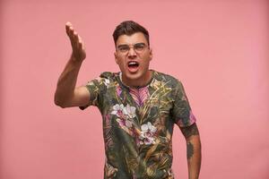 Close-up of young discontented man, showing to camera with his palm, frowning with widely opened mouth, standing over pink background photo