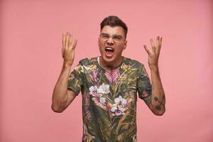 Uncomprehending puzzled young man with hands up standing over pink background, wearing t-shirt with floral print and glasses, looking confused photo