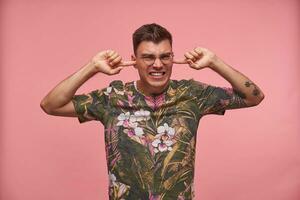 Stressed short haired young man with closed eyes pluging ears, avoiding loud sounds, ignoring something unpleasant, isolated over pink background photo