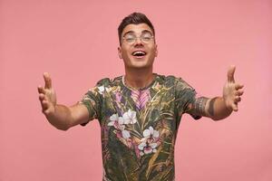 Pretty young man in flowered t-shirt smiling friendly, going to give a hug, standing over pink background with arms outstretched photo