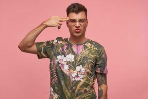 Sad short-haired young man curving lips and trying to kill himself, being tired of everything, shooting in temple with hand, isolated over pink studio background photo