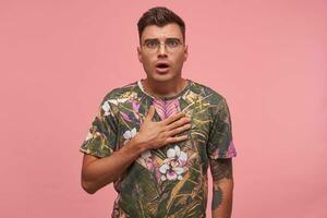 Headshot of pretty guy in flowered t-shirt, holding palm on his chest, feeling sorry, looking sad, standing over pink background photo