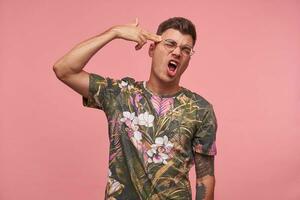 Young man with wide mouth open makes suicide gesture, being disappointed in everything, shoots in temple with hand, isolated over pink studio background photo