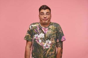 Indoor portrait of attractive guy standing over the pink background wih raised eyebrows, shrugging and making face, being bored and indifferent photo