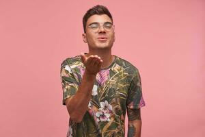 Close-up shot of handsome young guy with eyewear, expressing love, blowing kiss at camera, looking flirty, isolated over pink background photo