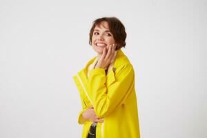 Portrait of young happy cute short haired girl wears in yellow rain coat, wbroadly smiles and looks at the camera, touches cheek, stands over white wall. photo