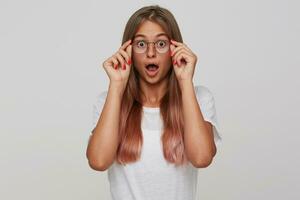 Studio shot of amazed young long haired blonde lady wearing glasses while standing over white background, rounding surprisedly her brown eyes while looking at camera photo