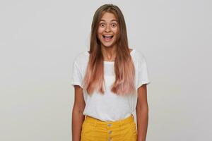 Amazed young attractive long haired blonde lady looking at camera with wide eyes and mouth opened, keeping hands along her body while standing over white background photo