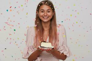 estudio Disparo de alegre joven encantador dama con ligero marrón largo pelo participación plato con cumpleaños pastel y mirando felizmente a cámara con amplio sonrisa, aislado terminado blanco antecedentes foto