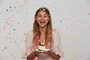 Overjoyed young long haired woman with casual hairstyle celebrating her birthday with friends, having fun on party and keeping plate with cake in raised hands while standing over white background photo