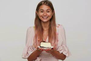 Pleasant looking young attractive brown-eyed long haired blonde lady smiling positively while keeping piece of cake in raised hands, standing over white background photo