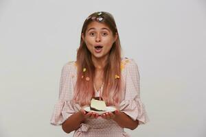 Surprised young pretty long haired blonde woman in festive elegant clothes looking excitedly to camera and holding piece of birthday cake, isolated over white background photo