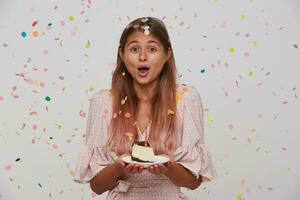 Studio shot of young beautiful long haired blonde lady celebrating her birthday with friends, standing over white background in paper confetti with cake in raised hands photo