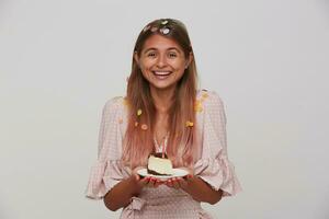 Positive young lady with light brown long hair smiling pleasantly at camera while rejoicing about birthday party with her friends, keeping piece of cake in hands while posing over white background photo