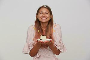 Pleased young beautiful long haired blonde female with charming smile posing over white background with piece of birthday cake, wearing festive elegant clothes photo