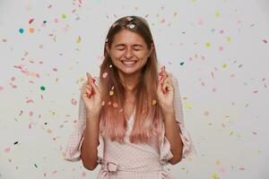 agradable mirando joven contento largo peludo rubia dama sonriente felizmente mientras haciendo deseo en su cumpleaños y acuerdo ojos cerrado, vistiendo rosado romántico vestir mientras en pie terminado blanco antecedentes foto