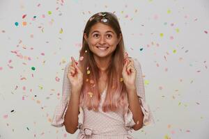 Positive young lovely brown-eyed long haired woman biting worringly underlip while making wish with crossed finfers, posing over white background in multicolored confetti photo