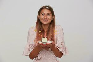 Portrait of good looking positive young long haired lady in festive clothes celebrating her birthday and standing over white background with piece of cake in raised hands photo