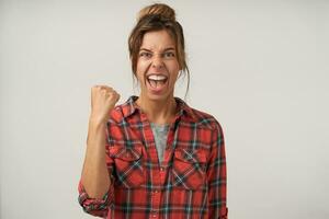 Excited young brown haired female with bun hairstyle raising fist in yes gesture while looking emotionally at camera with opened mouth, posing over white background photo