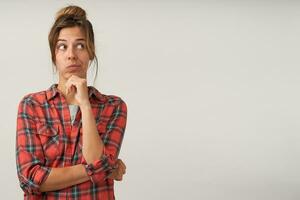 Puzzled young lovely brown haired female with natural makeup wrinkling forehead and holding chin with raised hand while looking pensively aside, isolated over white background photo