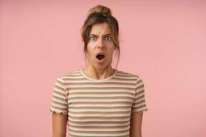 Confused young attractive brown haired woman keeping her mouth wide opened while looking indignantly at camera, posing over pink background in striped t-shirt photo