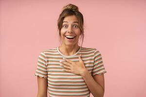 Excited young attractive brown haired lady raising surprisedly eyebrows while looking joyfully at camera and keeping palm on her chest, isolated over pink background photo