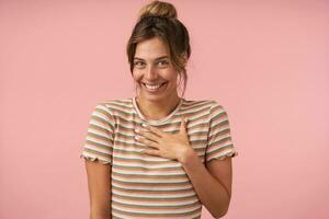 Portrait of young beautiful brown haired woman with natural makeup looking cheerfully at camera with wide smile while posing over pink background in casual wear photo