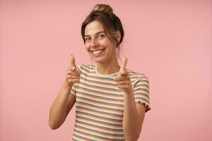 Indoor shot of young beautiful brown haired woman with natural makeup smiling happily while showing with raised hands at camera, isolated over pink background photo