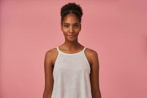 Portrait of young pretty brown-eyed brunette dark skinned woman keeping her lips folded while looking at camera with calm face, isolated over pink background photo