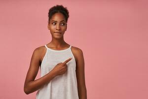 Puzzled young pretty curly brunette dark skinned woman with casual hairstyle showing aside with index finger and biting her undelip, isolated over pink background photo