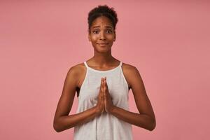 Hoping young lovely brown haired curly lady with dark skin keeping raised palms together and frowning eyebrows while looking at camera, isolated over pink background photo