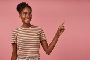 Studio photo of young glad brown haired curly lady with dark skin showing aside with forefinger and smiling broadly, posing over pink background in striped t-shirt