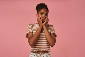 Portrait of afraid young pretty brown haired curly female with dark skin holding her face with raised hands and looking at camera with frightened face, posing over pink background photo