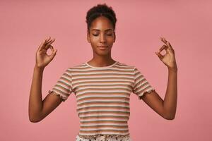 Pleasant looking young brown haired curly dark skinned lady raising hands with mudra gesture while meditating with closed eyes, isolated over pink background photo