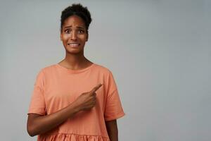 Indoor shot of confused young dark skinned curly female keeping her hand raised while showing aside with index finger and grimacing her face, standing over grey background photo