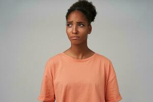 Portrait of upset young dark skinned curly lady with bun hairstyle frowning eyebrows and keeping lips folded while looking sadly aside, isolated over grey background photo