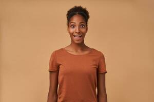 Agitated young lovely curly dark skinned lady with bun hairstyle raising amazedly eyebrows while looking excitedly at camera, standing over beige background photo