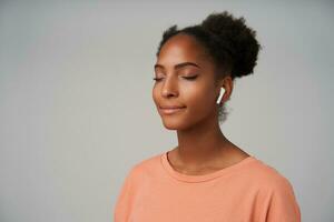 Pleasant looking young glad dark skinned curly woman with bun hairstyle smiling gladly with closed eyes while listening to music with earphones, isolated over grey background photo