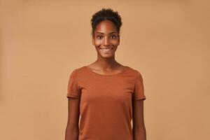 Indoor photo of young joyful brown haired curly woman with dark skin smiling cheerfully at camera while standing over beige background with hands down