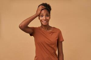 Bewildered young attractive brown-eyed dark skinned curly woman raising hand to her head while looking confusedly at camera, standing over beige background photo