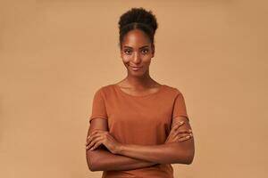 Indoor photo of young positive curly brunette lady with dark skin folding her hands on chest and smiling slightly at camera, standing over beige background