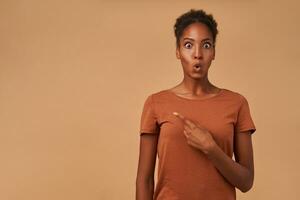 Dazed young attractive curly brunette female with dark skin rounding amazedly her eyes while showing aside with index finger, standing over beige background photo