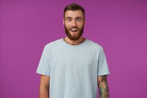 Amazed young bearded brunette with trendy haircut wearing blue t-shirt, looking at camera with wide eyes and mouth opened, posing over purple background with hands down photo